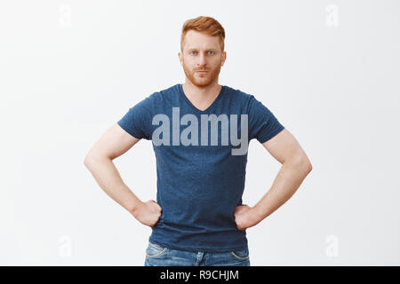 Tell me truth, I am listening. Serious strict and handsome redhead boyfriend in blue t-shirt, holding hands on waist and staring at camera, scolding someone or giving directions, being bossy Stock Photo