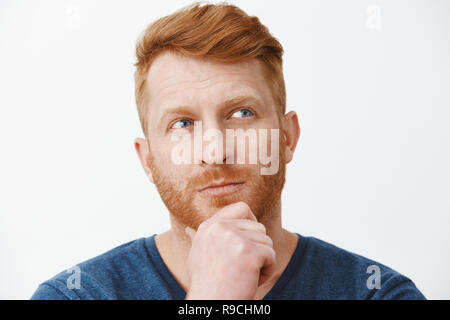 Headshot of thoughtful good-looking strategic man with bristle, touching chin and looking at upper right corner focused, thinking or making decision trying to pick right choice over gray wall Stock Photo