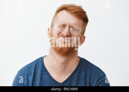 Close-up shot of cute and funny adult with red hair and beard, crying and being gloomy, closing eyes, frowning, tilting head back while pursing lips, feeling regret or upset over gray background Stock Photo