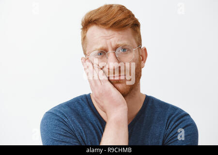 Headshot of upset and gloomy redhead guy with beard in transparent glasses, frowning holding palm on cheek gazing with regret and disappointment, losing chance over gray background Stock Photo