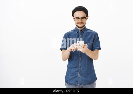 Confident smart and creative male studio manager in beanie and glasses with beard typing message in smartphone or searching information in internet gazing at gadget screen relaxed over gray wall Stock Photo