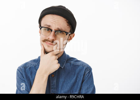 Close-up shot of doubtful smart suspicious handsome young guy with beard and moustache in black beanie and glasses touching chin and smirking squinting at camera feeling uncertain and unimpressed Stock Photo