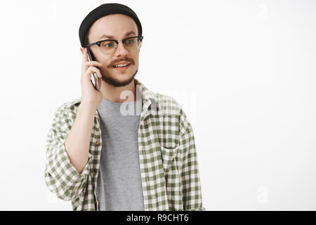 Waist-up shot of carefree calm and relaxed normal guy in beanie and glasses looking right with careless expression while calling friend talking on smartphone holding device near ear Stock Photo