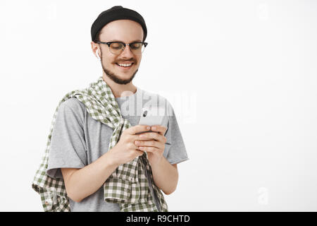 Joyful relaxed and delighted young boyfriend with beard in glasses and black hipster beanie holding smartphone smiling at device screen having funny and interesting conversation via internet Stock Photo