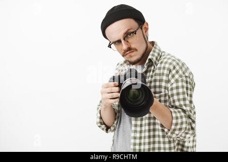 Waist-up shot of funny serious-looking male photographer in black beanie glasses and checked shirt pointing professional camera forward and gazing seriously to take picture during work Stock Photo
