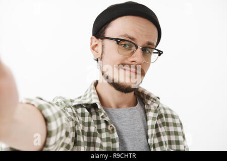 Close-up shot of flirty self-assured young handsome man with beard in black stylish beanie and checked shirt pulling hand towards camera smirking while taking selfie feeling confident in own look Stock Photo