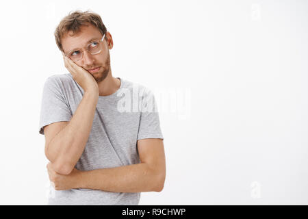 Waist-up shot of bored nostalgic gloomy guy in transparent glasses with bristle leaning head on palm staring at upper right corner with jealousy and sadness feeling boredom over white wall Stock Photo