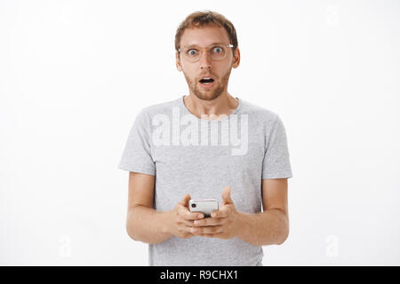Portrait of concerned shocked mature bearded male gasping opening mouth shocked holding smartphone staring at camera reading strange and disturbing message posing over white background Stock Photo