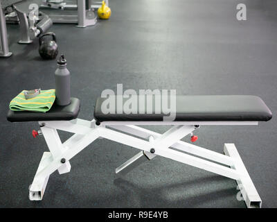 empty sport bench in gym with water bottle, smart watch and towel. high angle shot with dumbbells on dark floor blurred on background. Stock Photo