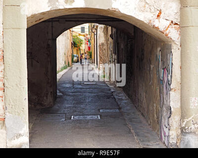 two men carry pack of bottled water below shuttered windows in typical narrow street with manhole covers and graffiti under archway in Pisa, Italy Stock Photo