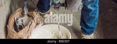 Young male painter kneads putty with water in a bucket using a hand-held mixer for building mixes BANNER, LONG FORMAT Stock Photo