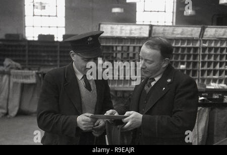 1948, picture shows two uniformed Royal Mail staff inside a postal sorting office, England, UK, a postman with cap standing with his supervisor checking the address on a letter. Stock Photo