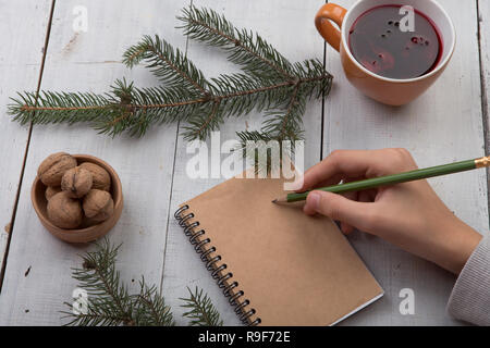 Winter holidays concept - blank notebook, pencil in hand, nuts, fir branches and cup of tea on white wooden table Stock Photo