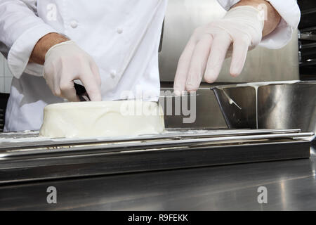 hands pastry chef prepares a cake, cover pouring white icing, working on a stainless steel industrial kitchen work top Stock Photo