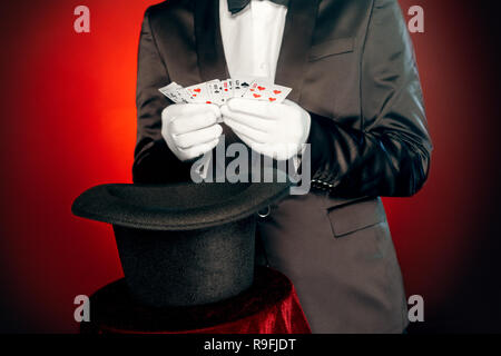 Professional magician wearing suit and gloves standing isolated on black and red background showing tricks with cards and top hat close-up Stock Photo