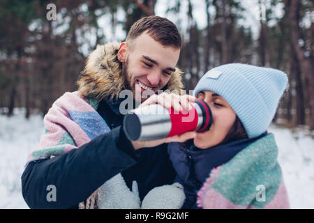 Man giving his girlfriend hot tea to drink in thermos cup. Loving couple walking in winter forest together covered with blanket Stock Photo