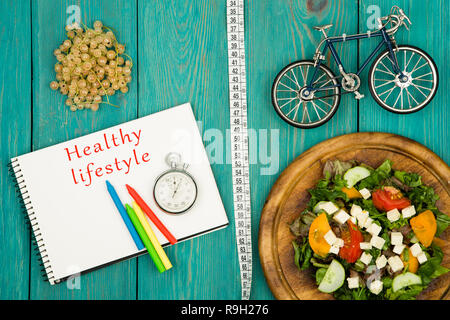 bicycle model, salad of fresh vegetables, notepad with text "Healthy lifestyle", stopwatch and tape measure on a blue wooden table Stock Photo