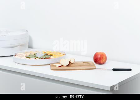On a white tabletop there is a red apple, a cutting board, a kitchen knife, a dehydrator and a tray with slices of fruit. Stock Photo