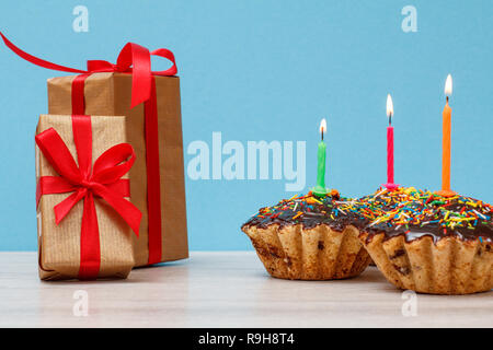 Gift boxes and three tasty birthday cupcakes with chocolate glaze and caramel, decorated with burning festive candles on blue background. Happy birthd Stock Photo