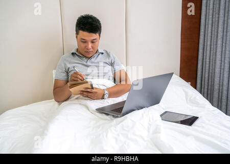 Young Asian man working and writing notes on bed Stock Photo