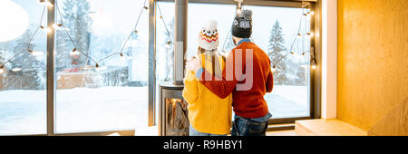 Young couple dressed in bright sweaters enjoying landscape view on the mountains standing together in the modern house during winter time Stock Photo