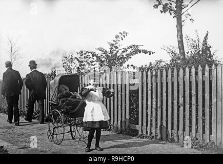 A late Victorian black and white photograph showing a very young girl with a pram with a bay in it, on a street in England, while two men walk away behind the pram. The pram has large iron wheels, and all the figures show good examples of fashions of the era. Stock Photo