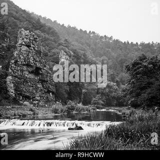 A late Victorian black and white photograph showing Dovedale, a valley in the Peak District of England. The land is now owned by the National Trust, and annually attracts a million visitors The valley was cut by the River Dove and runs for just over 3 miles (5 km) between Milldale in the north and a wooded ravine near Thorpe Cloud and Bunster Hill in the south. In the wooded ravine, a set of stepping stones cross the river, and there are two caves known as the Dove Holes. Stock Photo