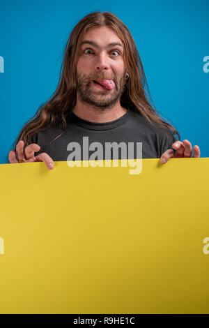 Male portrait making funny grimace while holding a yellow advertise banner. Long haired portrait of adult while holding sign with copy space available Stock Photo
