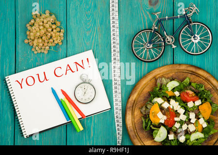 Diet and sport concept - bicycle model, salad of fresh vegetables, notepad with text "YOU CAN!", stopwatch and tape measure on a blue wooden table Stock Photo