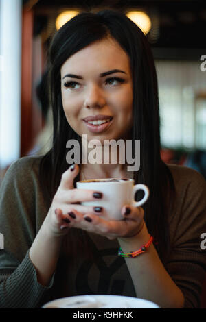beautiful girl with dimples on the cheeks in a cozy cafe against the window and bokeh. attractive model with the cup of coffee looks out the window Stock Photo
