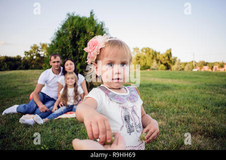 portrait of a beautiful little girl who gives the operator a grape. happy child in the background of her parents in the park Stock Photo