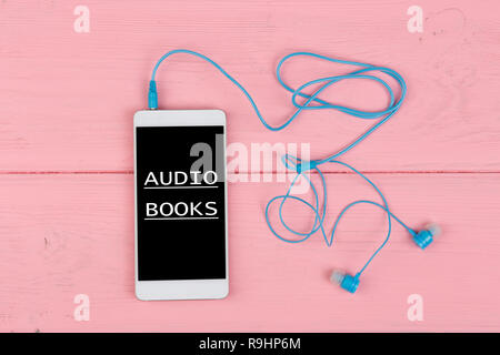 White smart phone with touch screen and text "Audio books", blue earphones on pink wooden table Stock Photo