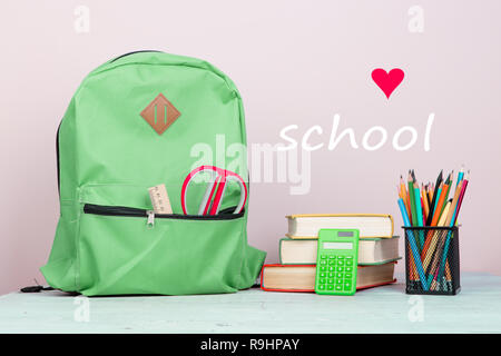 Green backpack and school supplies: notepad, books, scissors, pens, pencils, ruler, calculator is on a white wooden table on pink background with text Stock Photo