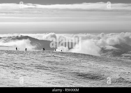 Black and white silhouettes of skiers and snowboarders on snowy ski slope and mountains in beautiful clouds at winter evening. Caucasus Mountains, Geo Stock Photo