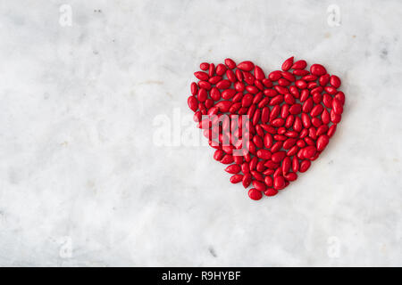 Red candy coated sunflower seeds in a heart shape on a white granite background for Valentine’s Day Stock Photo