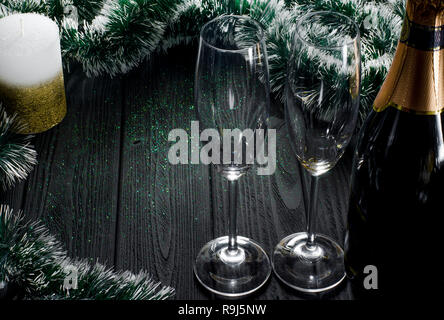 A bottle of champagne and two glasses stand on a dark gray wooden table surrounded by Christmas decorations and a white-gold candle stands next to it. Stock Photo