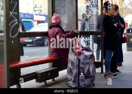 A woman with a shopping trolley sitting at a London bus stop, waiting for a bus Stock Photo