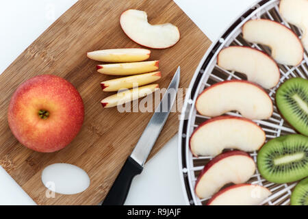 Cutting board with a knife and pieces of apples. Nearby is a dehydrator with slices of orange, kiwi and apples. Stock Photo