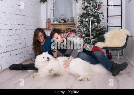 young beautiful couple having fun with their dog on the background of the New Year and Christmas decor. Family with Samoyed celebrate Stock Photo