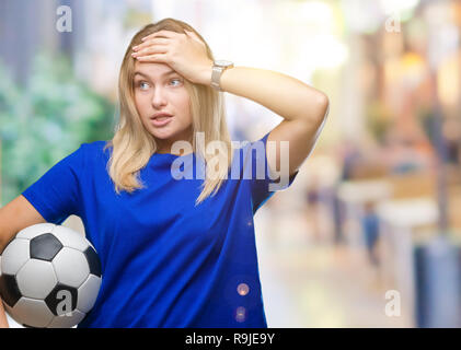 Young caucasian woman holding soccer ball over isolated background stressed with hand on head, shocked with shame and surprise face, angry and frustra Stock Photo