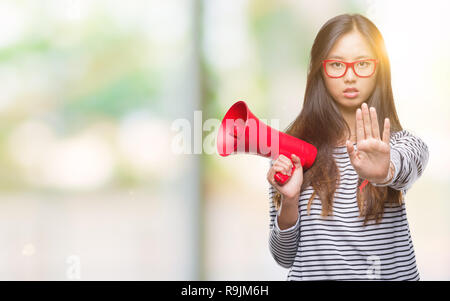 Young asian woman holding megaphone over isolated background with open hand doing stop sign with serious and confident expression, defense gesture Stock Photo