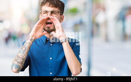 Young handsome man wearing glasses over isolated background Shouting angry out loud with hands over mouth Stock Photo
