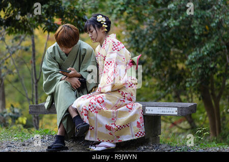 Japanese couple wearing traditional clothes sitting on wooden bench in a park, looking at phone, Kyoto, Japan Stock Photo