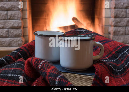 Two white enameled mugs with tea, and warm red wool scarf , a book , near cozy fireplace, in country house, winter vacation. Stock Photo
