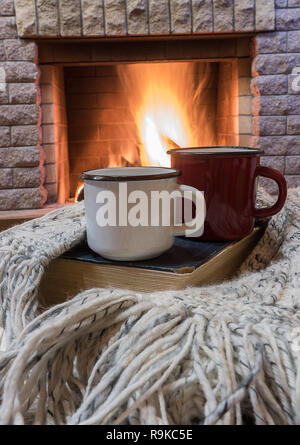 Big enameled mugs with tea, and warm wool scarf , opened book , near cozy fireplace, in country house, winter vacation, vertical. Stock Photo