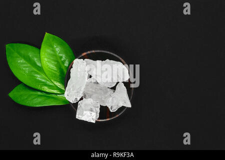 Abstract composition of green leaves, wooden dark bowl and big sugar or salt crystals on black background with copy space Stock Photo