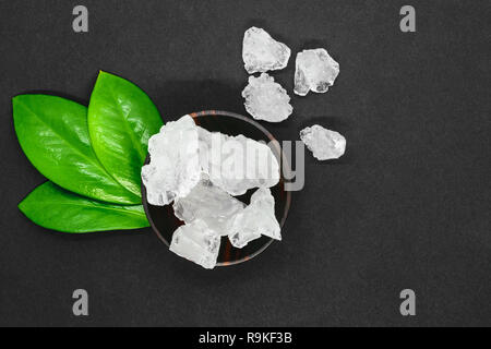Abstract composition of green leaves, wooden dark bowl and big sugar or salt crystals on grey background with copy space Stock Photo