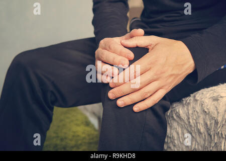 elderly man sitting on a sofa in the living room at home and touching his knee by the pain. man suffering from knee pain sitting sofa. Stock Photo
