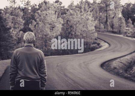 Active senior man stands alone on lonely road between mountains in black and white. older man of back walking on lonely highway for the mountains Stock Photo