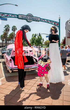 Costumed Elvis Presley impersonators of both sexes on stilts shake hands with a fan behind a pink Cadillac at an outdoor music festival in Fullerton, CA. Stock Photo